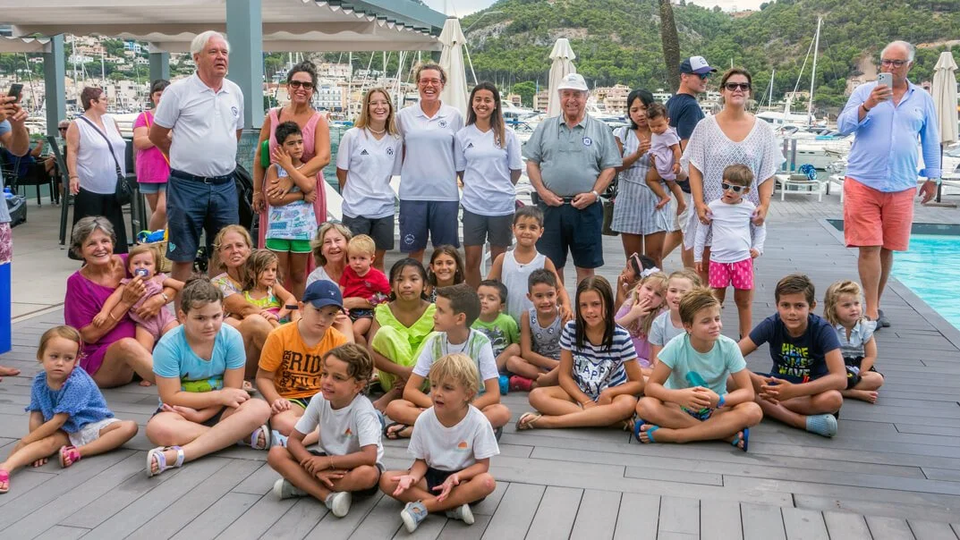 Grupo de personas, adultos y niños, posando frente a un muelle con yates, en un entorno con montañas y vegetación.