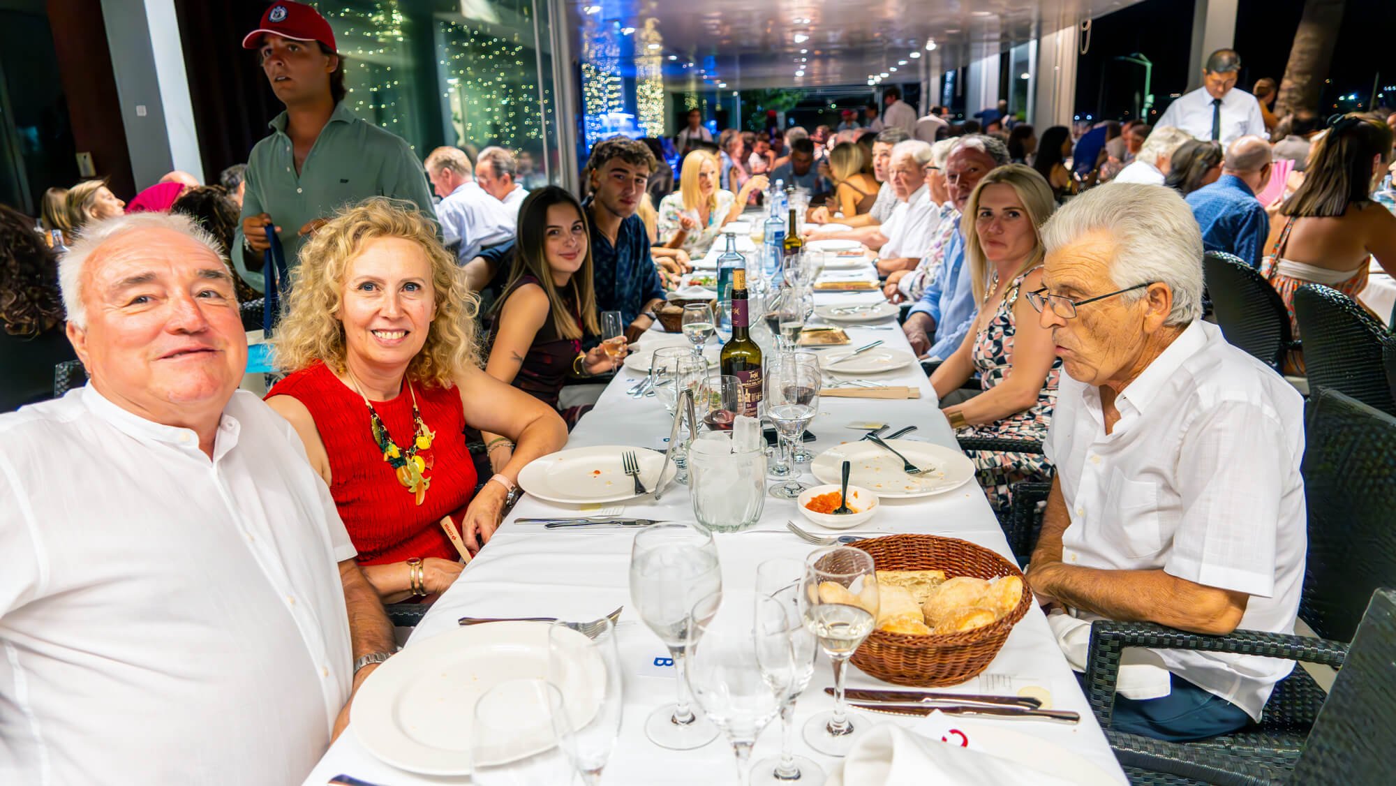 Grupo de personas sentadas en una mesa larga en un restaurante, disfrutando de una comida juntos por la noche, con decoraciones de luces y plantas alrededor.