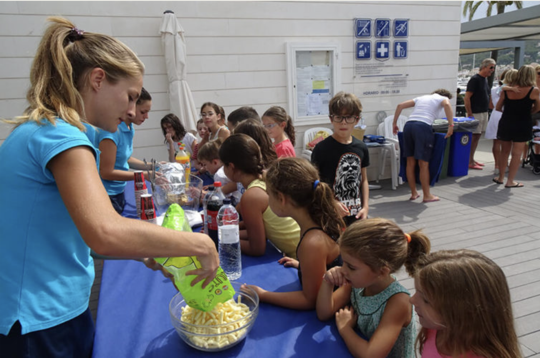 Niños y niñas en una mesa de evento al aire libre, esperando su comida, con adultos en el fondo y señales de indicaciones y primeros auxilios en la pared detrás.