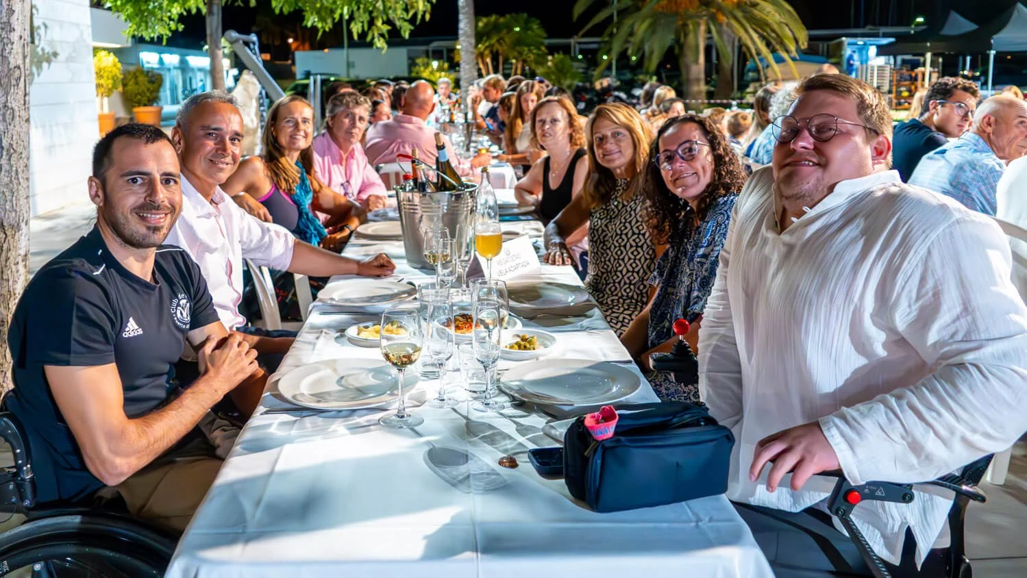 Grupo de personas en una comida al aire libre en la noche, sentadas en una mesa larga con platos, copas y botellas, algunos sonriendo y otros conversando.