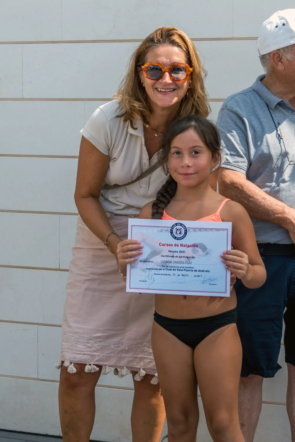 Una niña con traje de baño sosteniendo un certificado de natación, acompañada de una mujer con gafas naranjas. La niña tiene cabello castaño oscuro en una trenza y la mujer tiene cabello rizado y rubio. Están al aire libre, en un entorno soleado.
