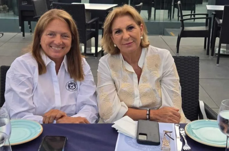 Dos mujeres sentadas en una mesa en un restaurante, una con blusa blanca con logotipo y otra con blusa beige, ambas sonriendo.