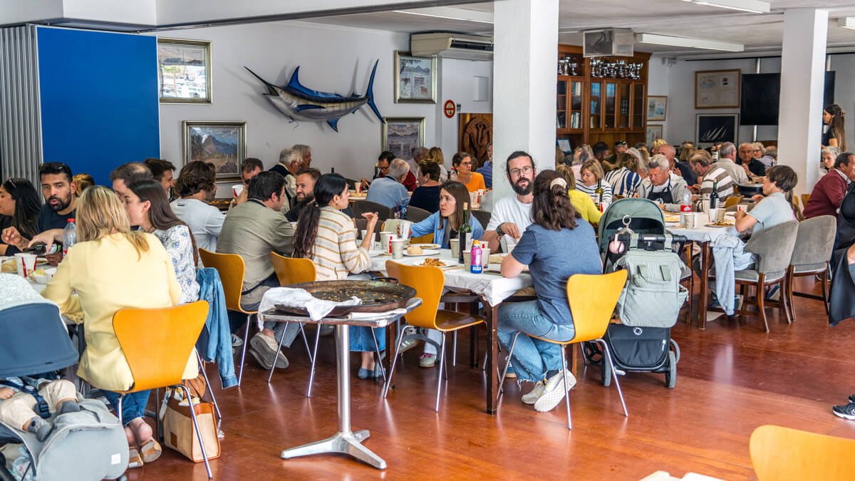 Muchas personas sentadas en mesas en un restaurante, algunos comiendo y otros conversando, con decoraciones marinas en la pared, incluyendo un pez espada y cuadros.