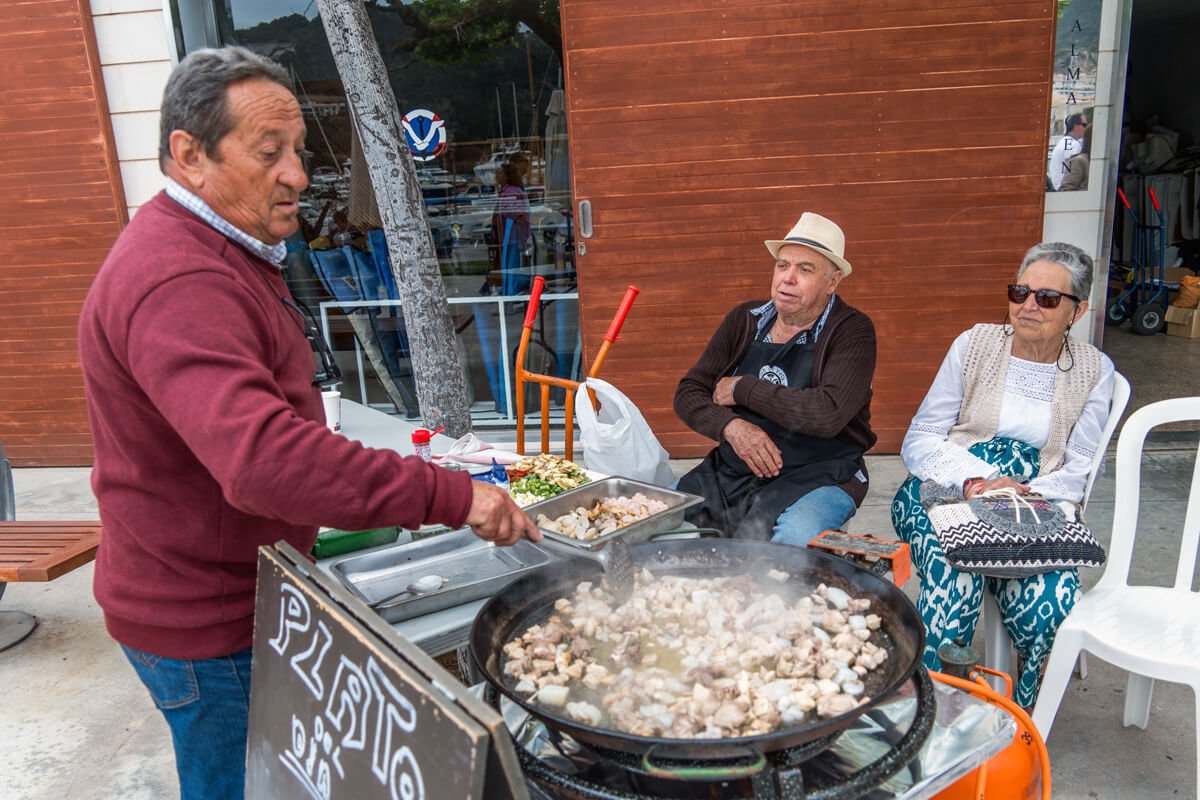 Un hombre cocina mariscos en una olla grande en una calle, mientras dos personas observan y esperan, una de ellas con gafas de sol y otra con sombrero, en un ambiente exterior.