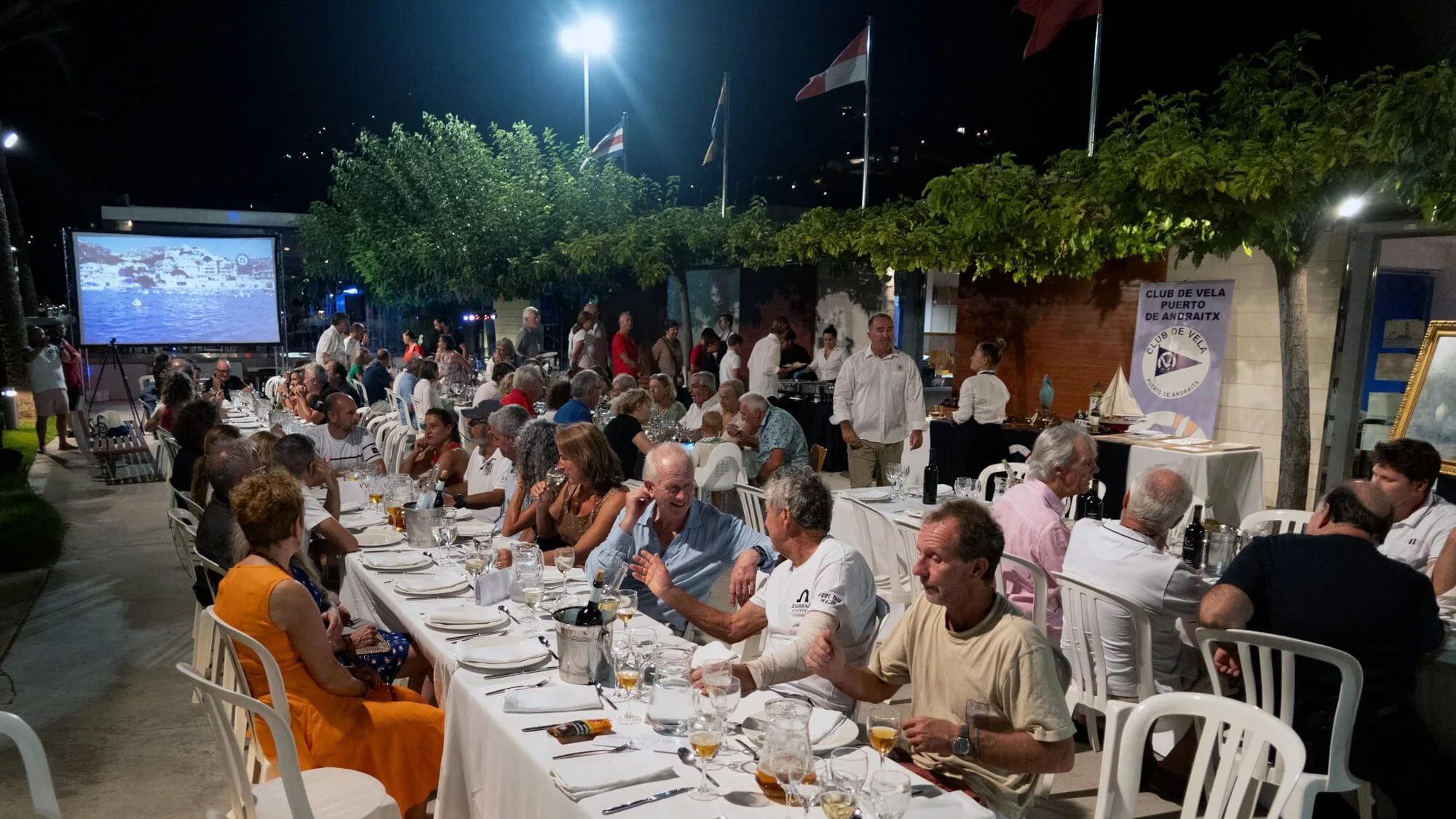 Cena de una cena al aire libre con muchas personas sentadas en mesas, algunos con comida y bebida, en un evento nocturno con banderas y una pantalla gigante en el fondo.