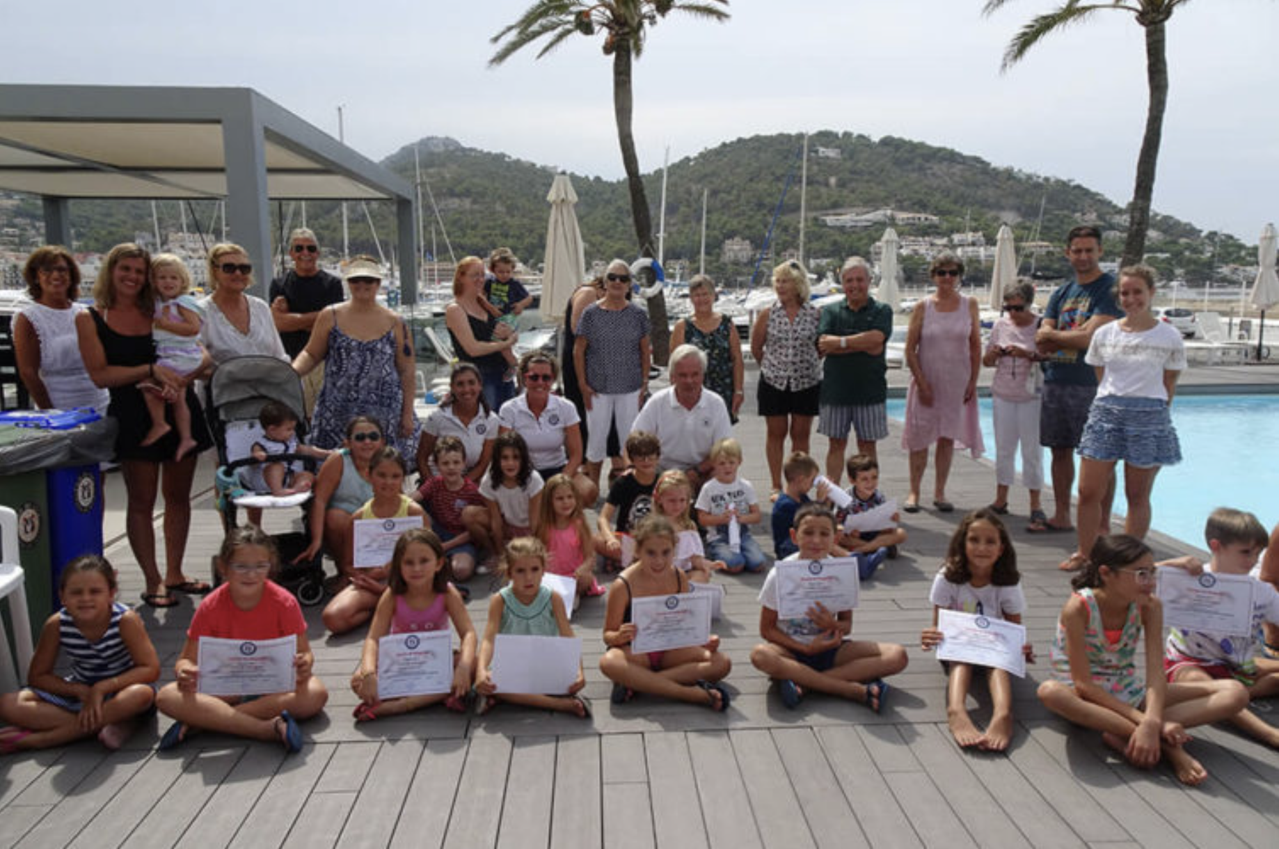 Grupo de personas, niños y adultos, en un muelle junto a un puerto con yates, en un día soleado y con palmeras, participando en un evento o ceremonia de entrega de certificados.