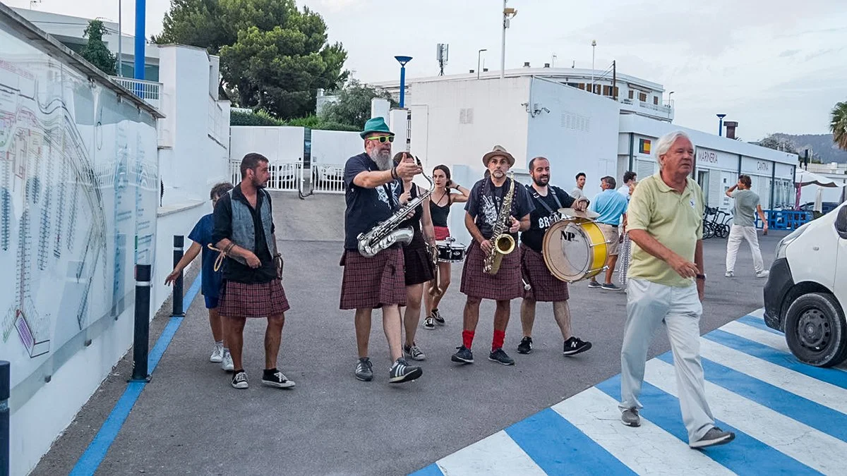Grupo de personas tocando instrumentos musicales en la calle, con un hombre mayor caminando frente a ellos, algunos niños y adultos en el fondo, en un área urbanizada con edificios y árboles.