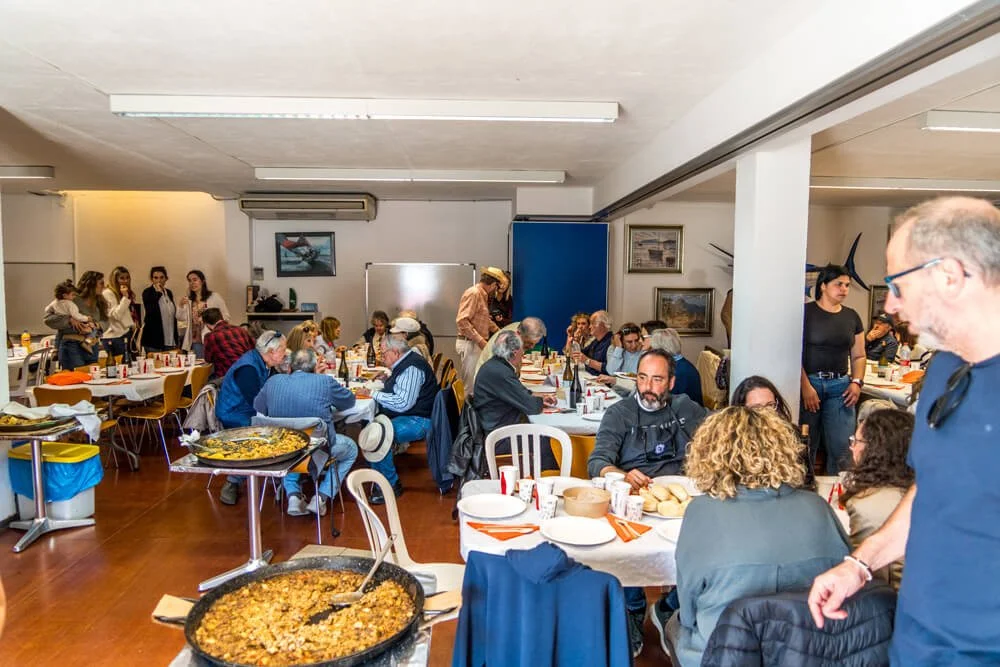Personas en una comida comunitaria en un salón, algunas sirviéndose y otras conversando, con comida y bebidas en las mesas.