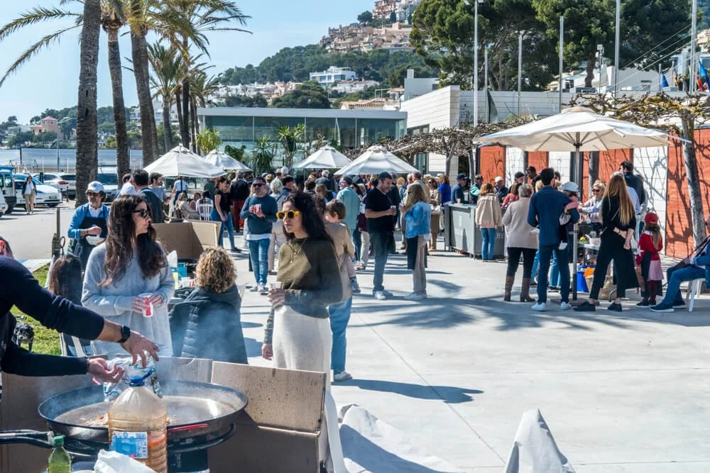 Personas en un evento al aire libre junto al agua, con sombrillas y árboles, disfrutando de comida y socializando.