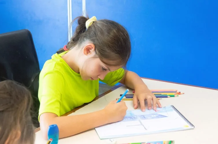 Una niña en una mesa de estudio, dibujando en un cuaderno, rodeada de lápices de colores.