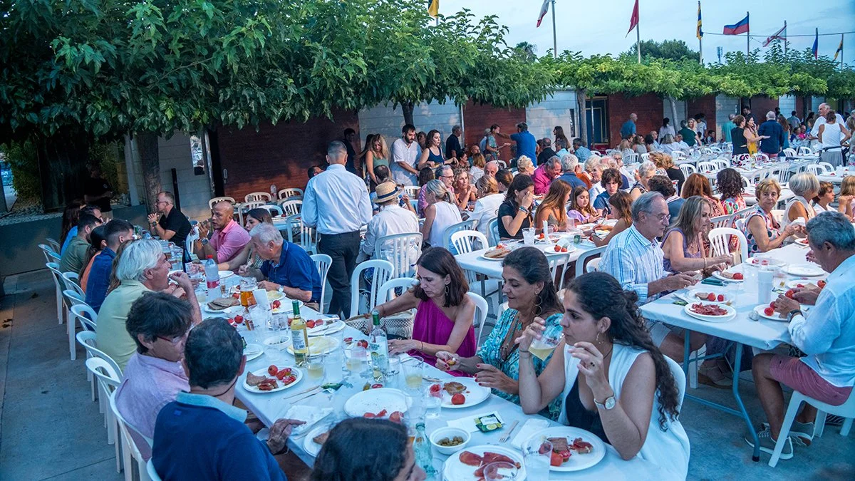 Una celebración al aire libre con muchas personas sentadas en mesas con comida, vegetación y banderas en el fondo.