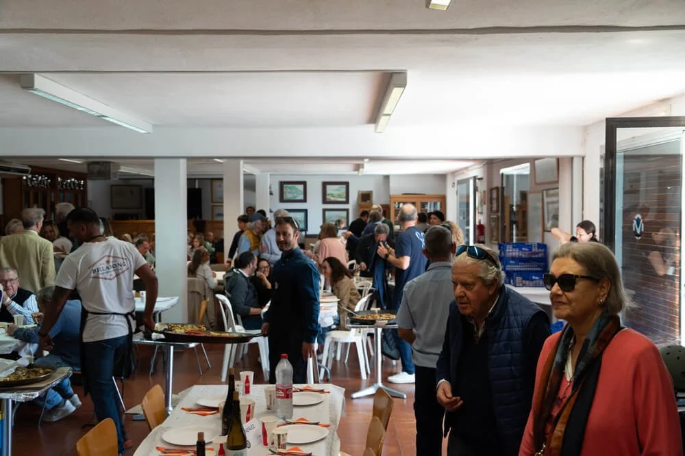 Imagen de una reunión o evento en un restaurante o salón, con muchas personas sentadas y de pie, conversando y disfrutando de comida y bebida.
