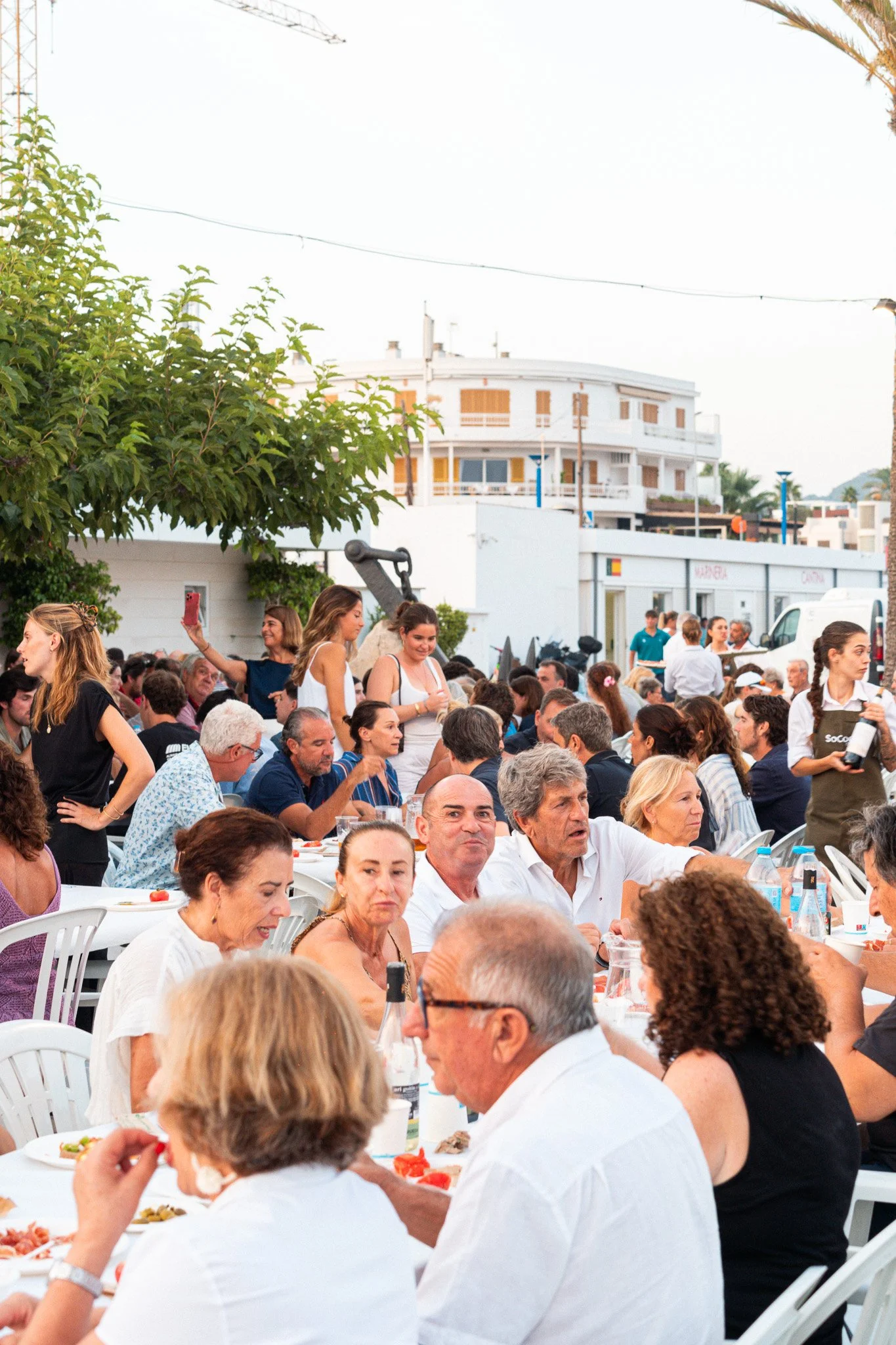 Personas sentadas en mesas blancas al aire libre, disfrutando de una comida, con árboles y edificios en el fondo.