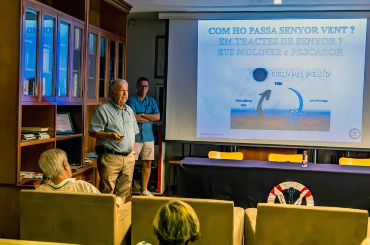 Una presentación en una sala con varias personas. En la pantalla hay un mapa del viento y un texto en español sobre cómo pasa el viento en verano.