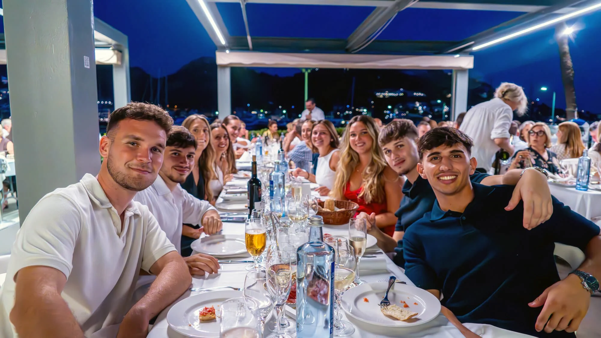 Grupo de personas en una cena en un restaurante al aire libre, sonrientes y brindando con comida y bebidas en la mesa, fondo con paisaje nocturno y luces.