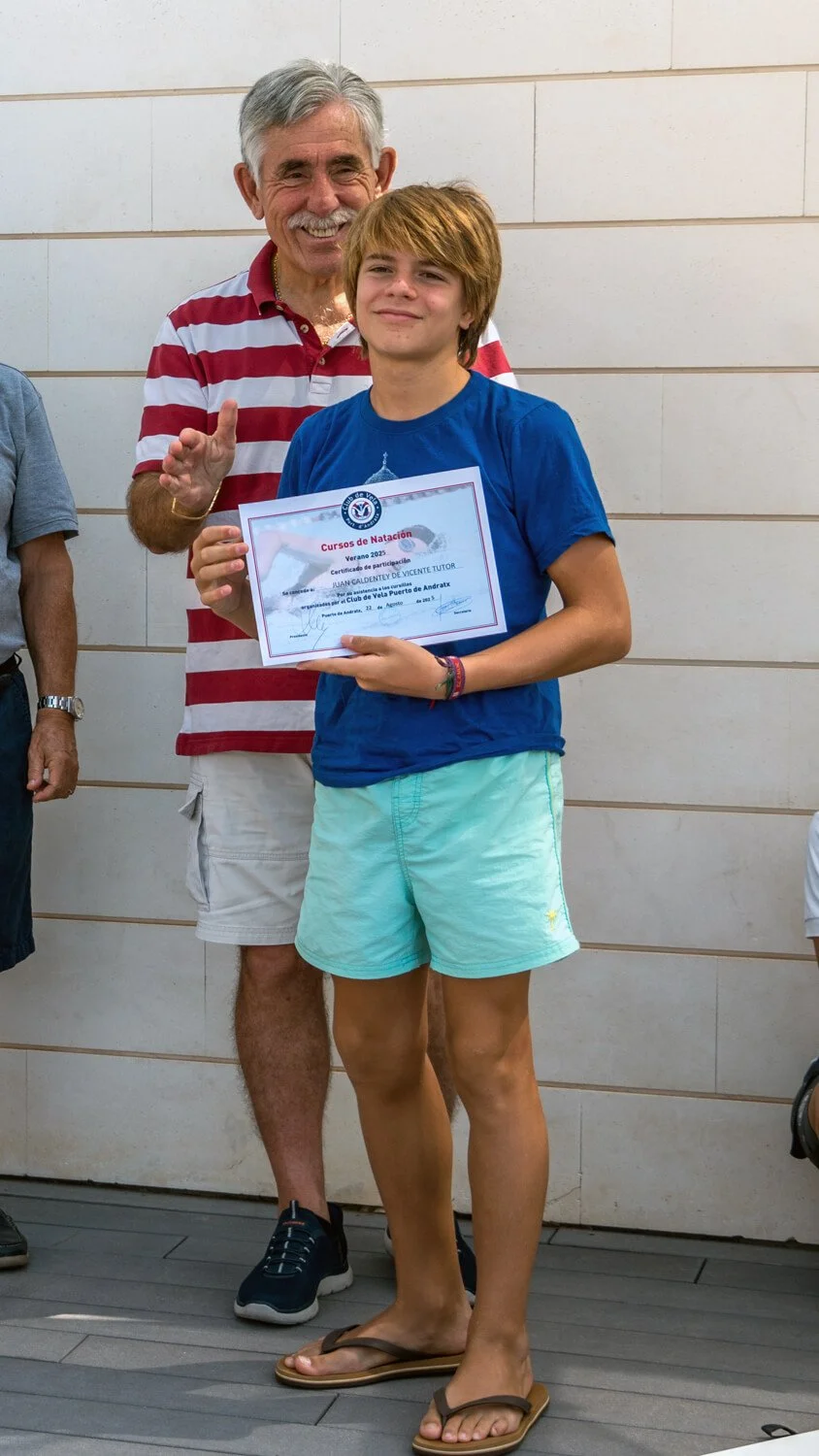 Un joven sostiene un certificado de participación en un curso de natación, mientras un hombre mayor le felicita con una sonrisa en su rostro, en un fondo de pared de azulejos.