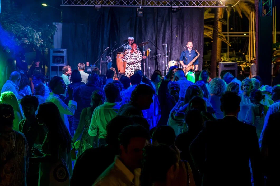 Concierto en un evento nocturno con público disfrutando música en un espacio abierto, con músicos en un escenario iluminado con luces de colores.