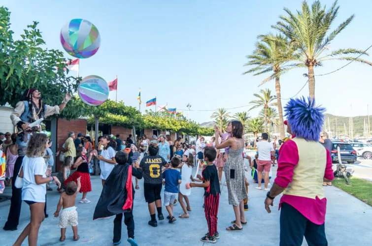 Personaje con pelo azul en un evento al aire libre, con niños y adultos bailando y disfrutando, palmeras y coches en el fondo.