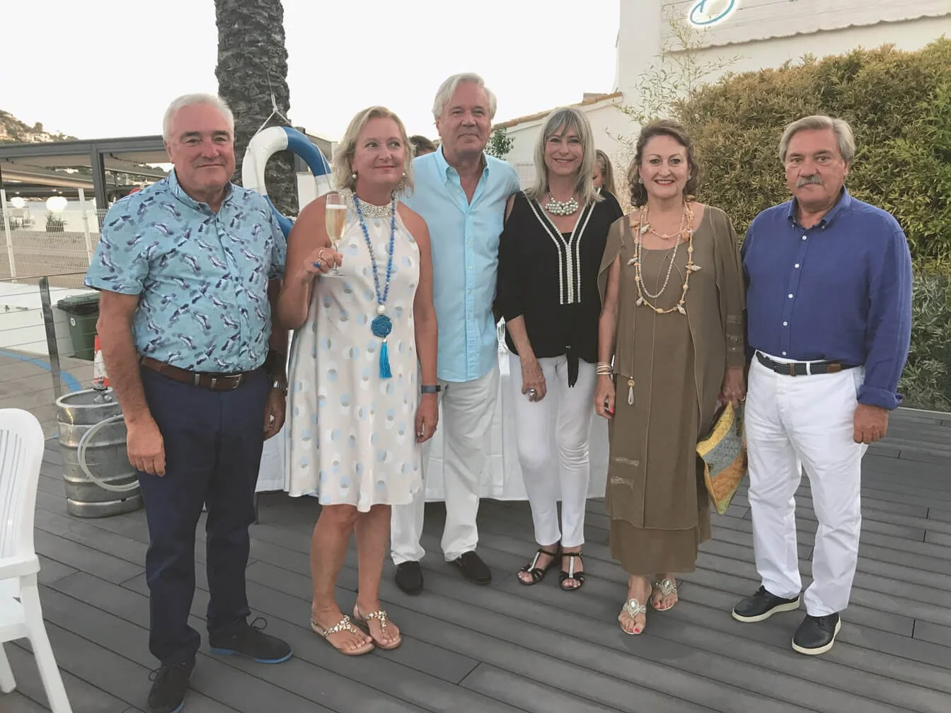 Grupo de seis personas en attire formal y casual en lo que parece ser una celebración o evento social al aire libre, con plantas y estructura en el fondo.
