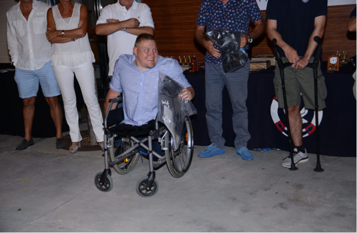 Hombre en silla de ruedas sonriendo rodeado de varias personas, algunas con brazos cruzados, en un ambiente interior con madera en el fondo.