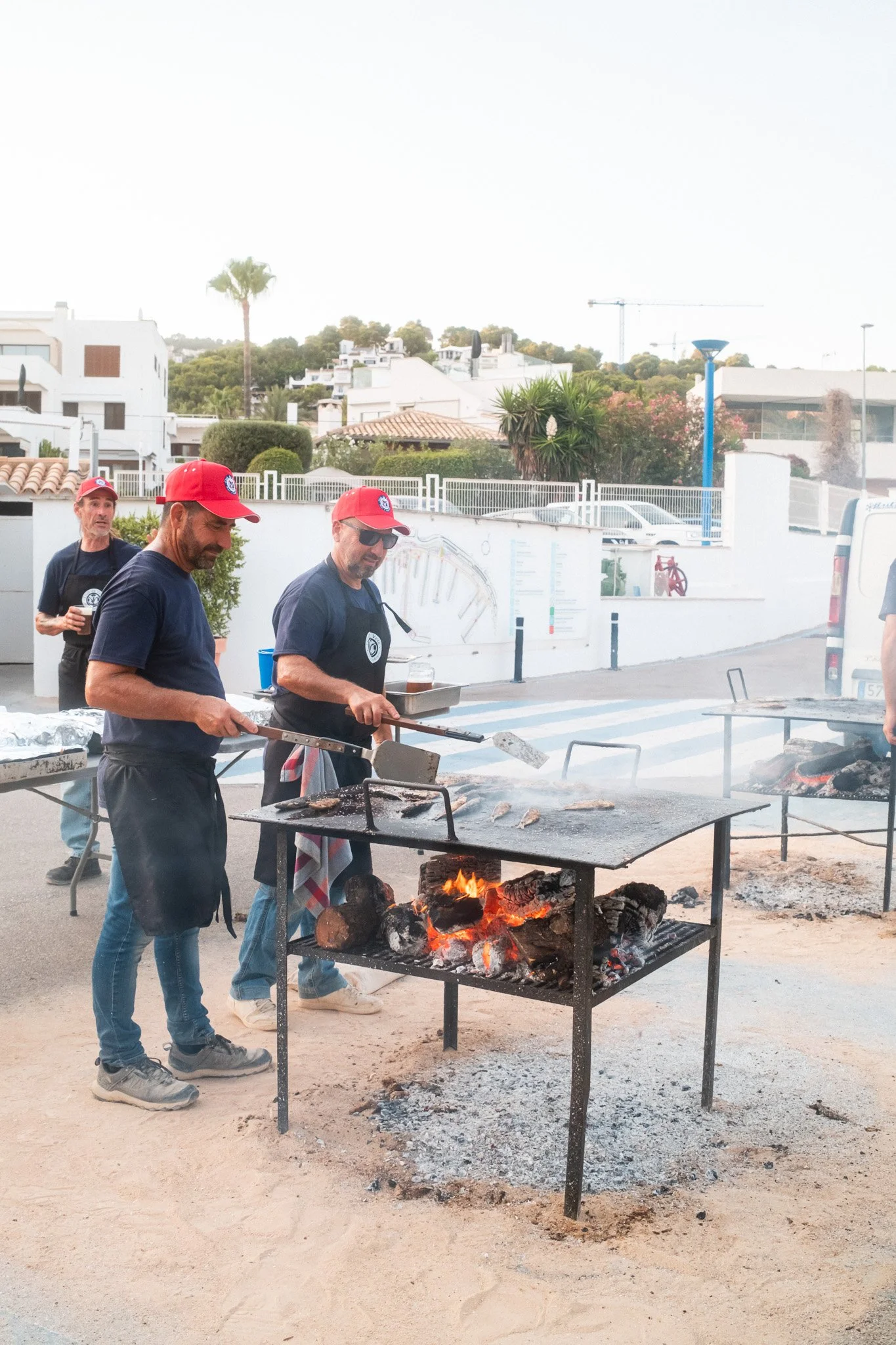 Hombres cocinando carne sobre una parrilla al aire libre con fuego de leña, en un entorno urbano con árboles y casas en el fondo.