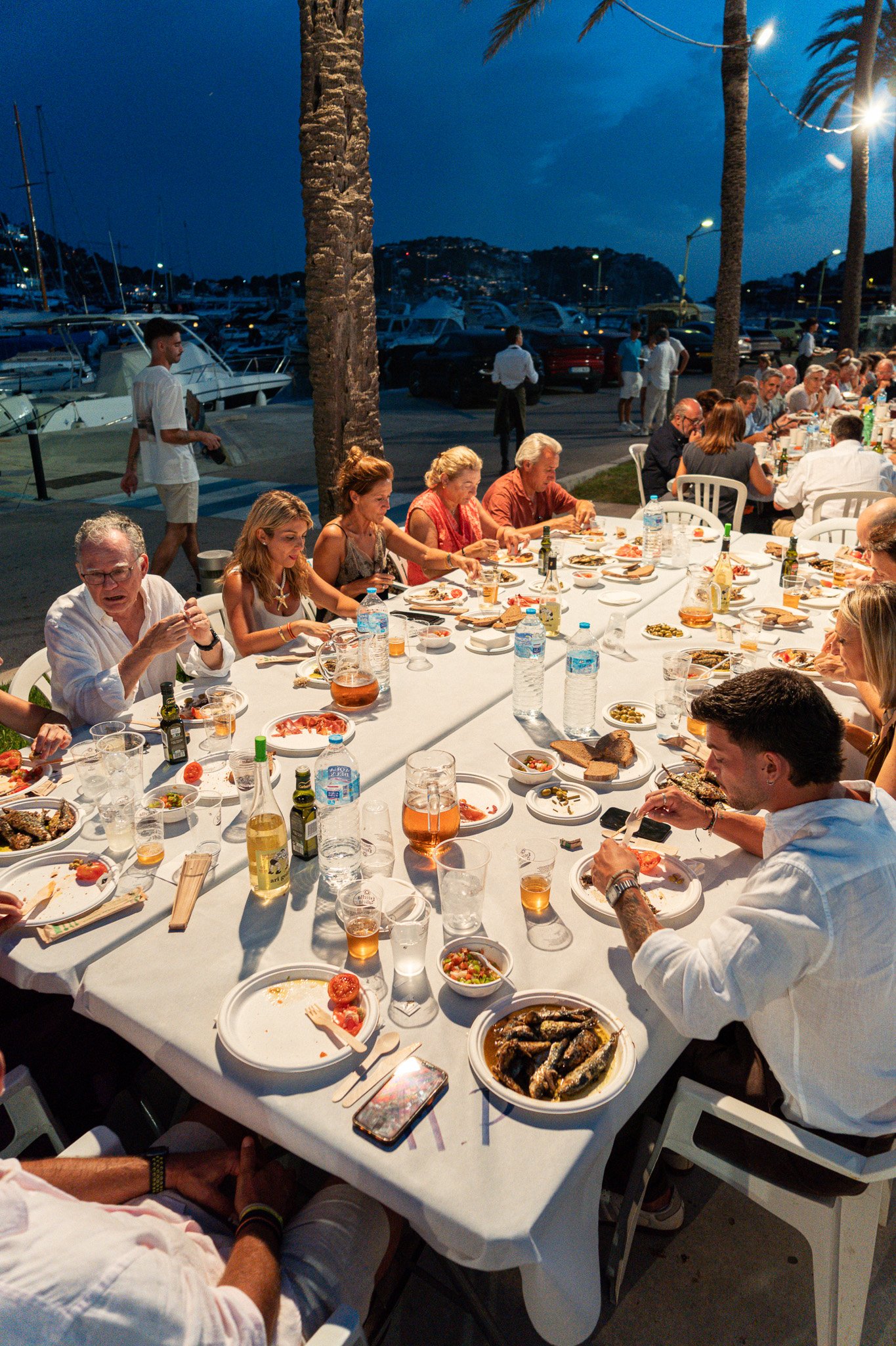 Personas cenando en una mesa larga al aire libre junto a un muelle con yates y coches, con palmeras y montañas de fondo, durante la noche.