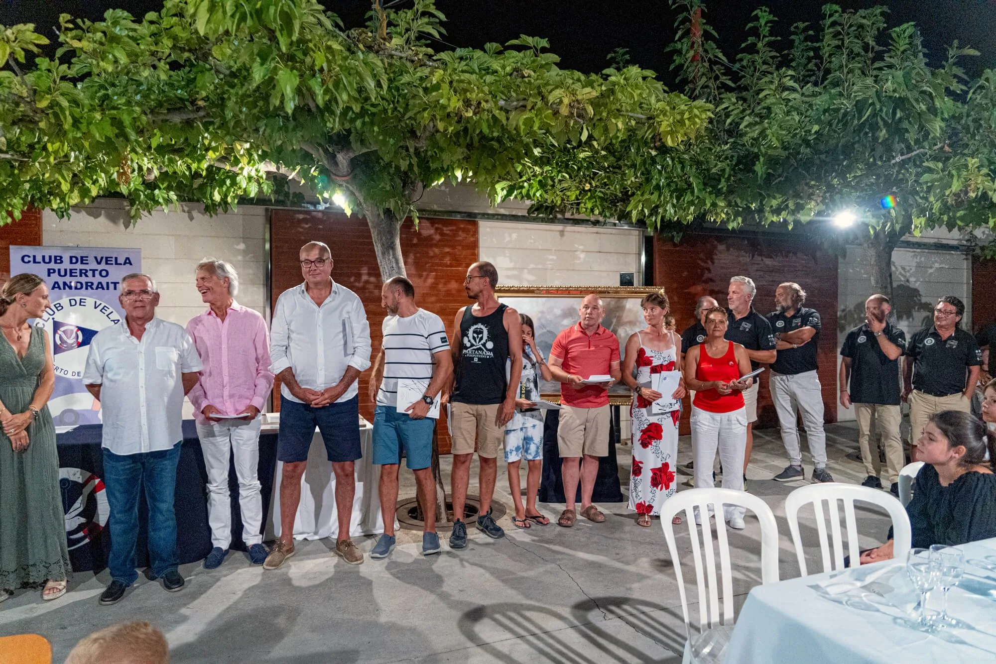 Grupo de personas en una reunión o evento al aire libre en la noche, con varias mujeres y hombres en una fila, algunos con documentos en las manos, y un cartel que dice "Club de Vela Puerto Andrait" al fondo.