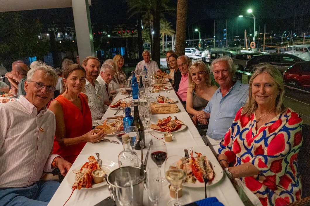 Grupo de personas disfrutando de una cena al aire libre en la noche, en una mesa larga con comida y bebida, junto a un muelle con barcos y palmeras en el fondo.