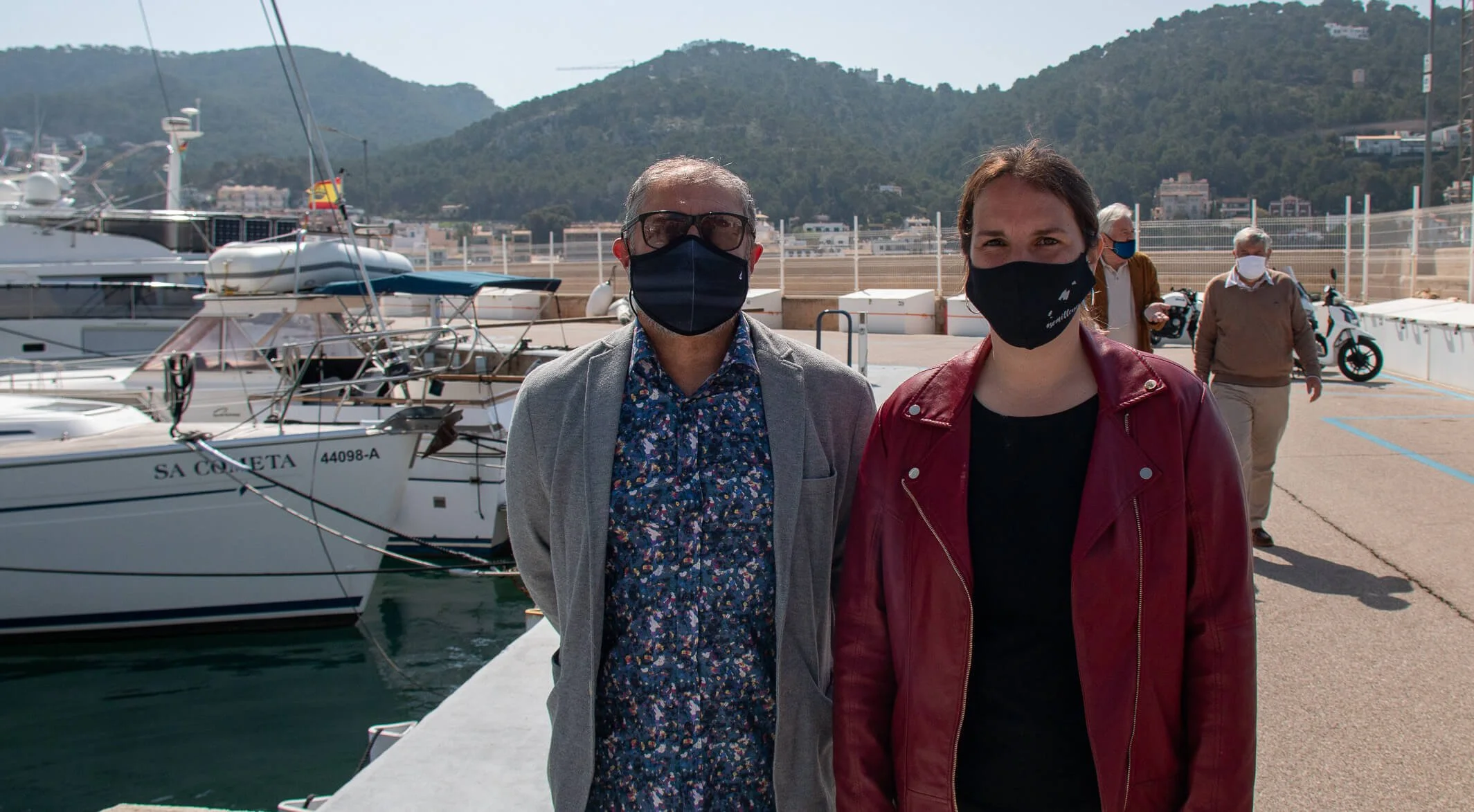 Dos personas con máscaras faciales enfrente de yates en un puerto, con montañas y edificios de fondo.