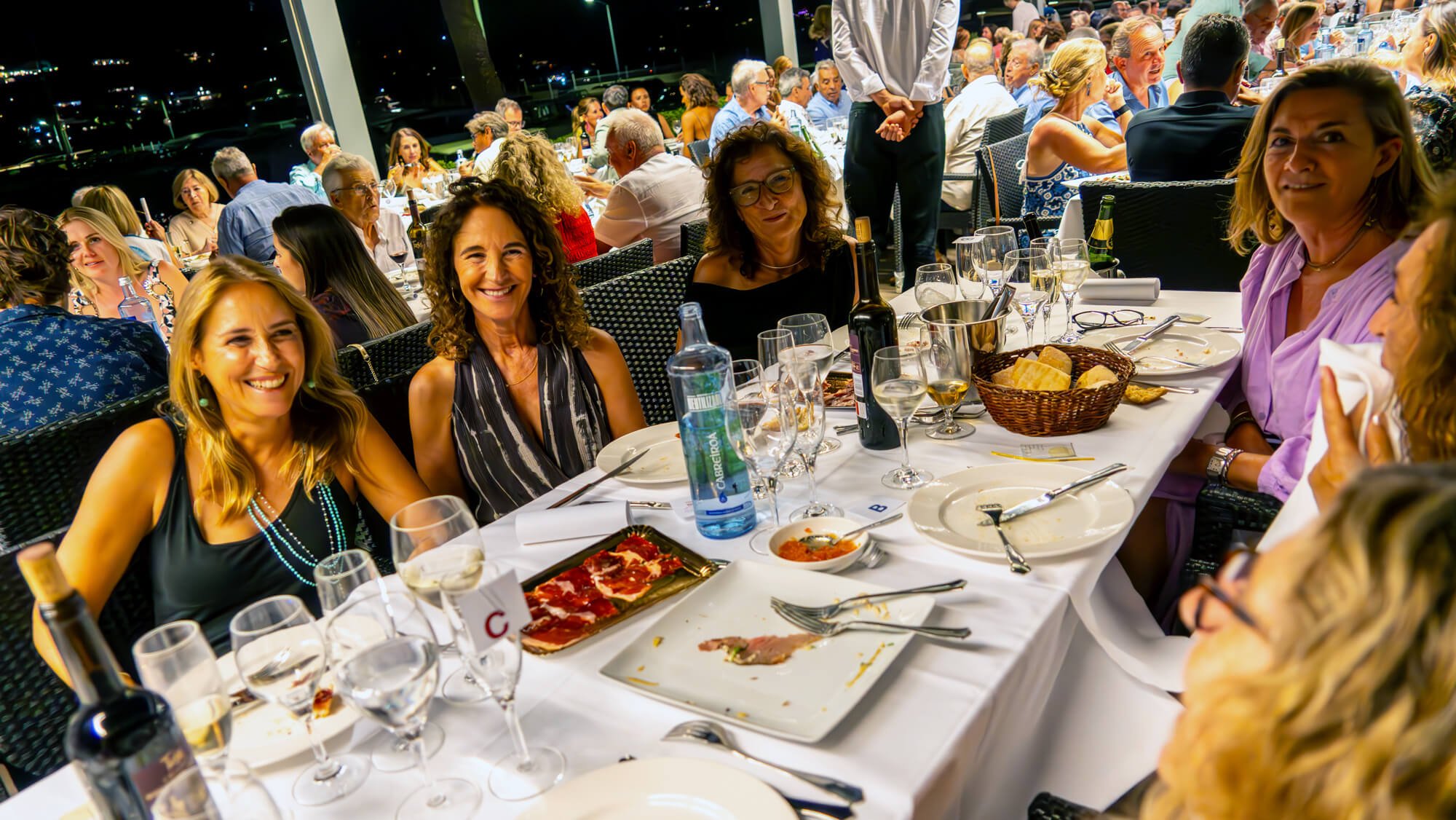 Cena en un restaurante con muchas personas, varias de ellas sonriendo y charlando, en una mesa con comida, vino y agua, en un ambiente nocturno con ventanas que muestran un paisaje de noche.