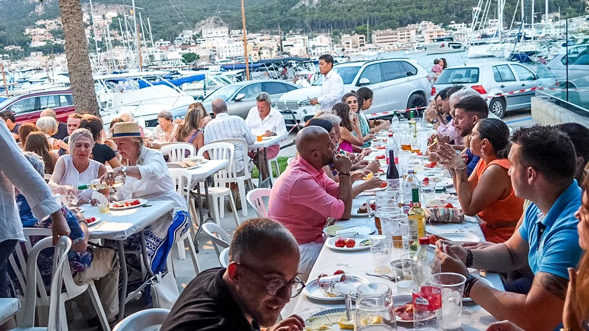 Reunión social al aire libre en un restaurante cerca de un puerto con yates, con muchas personas sentadas en mesas largas disfrutando de comida y bebida.