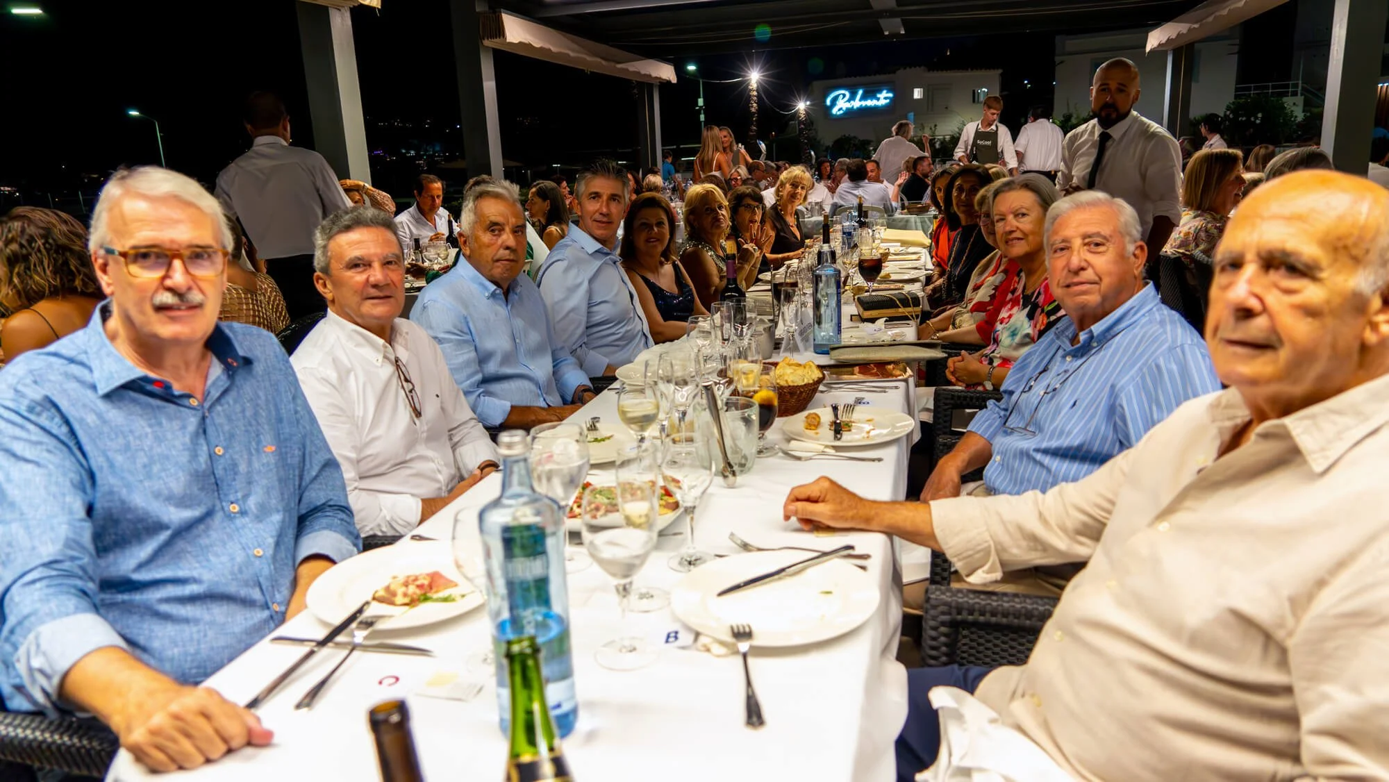 Grupo de personas en una cena elegante en un restaurante durante la noche, con platos de comida y bebidas sobre la mesa.