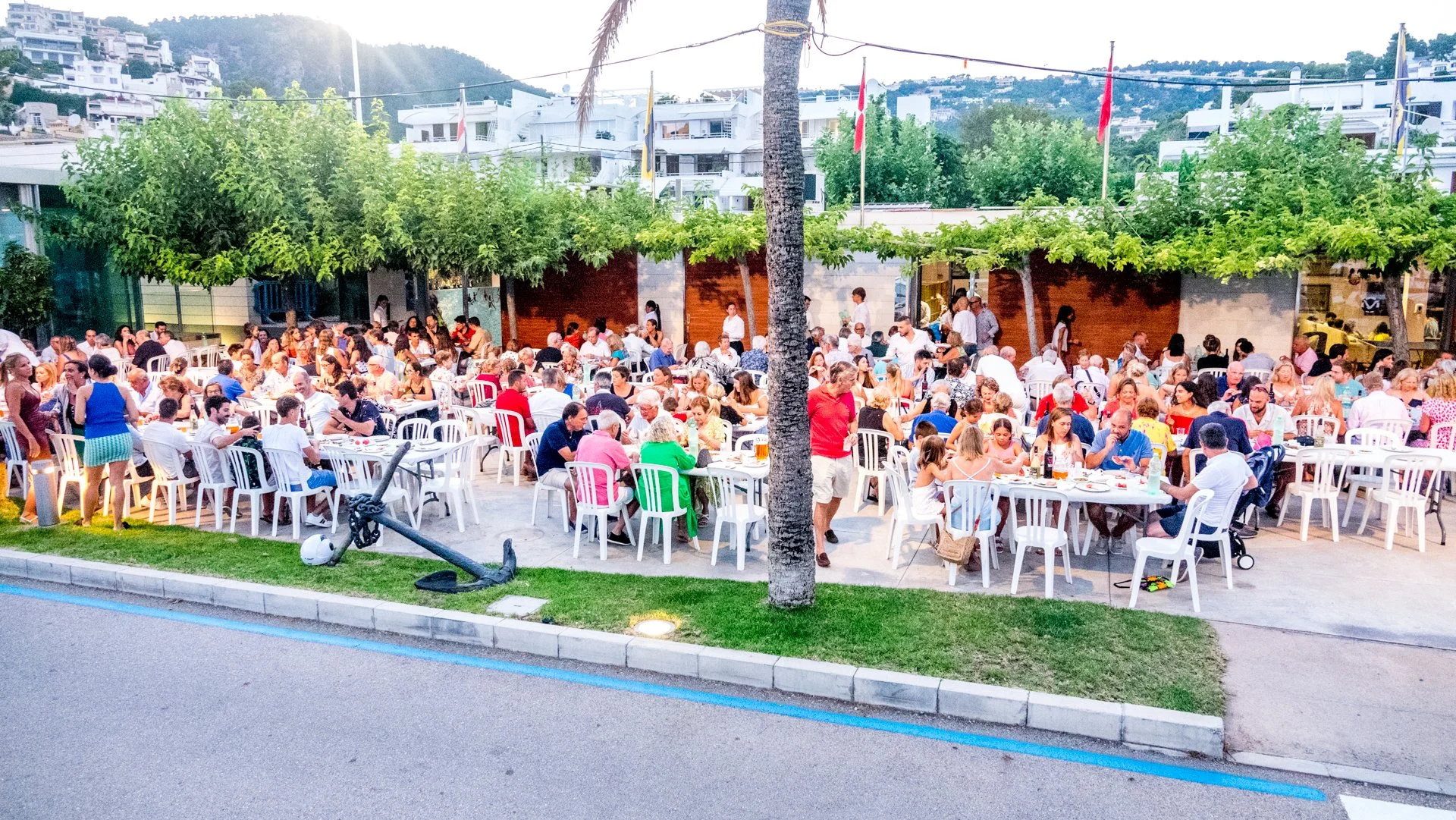 Una reunión grande al aire libre con muchas personas sentadas en mesas blancas en un espacio decorado con árboles y banderas, en un entorno urbano con colinas y edificios blancos en el fondo.