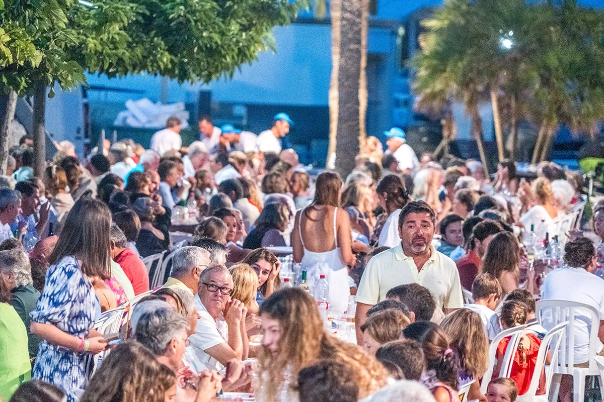 Una gran reunión al aire libre con muchas personas sentadas en mesas, probablemente en una fiesta o evento social, con árboles y un ambiente vespertino.