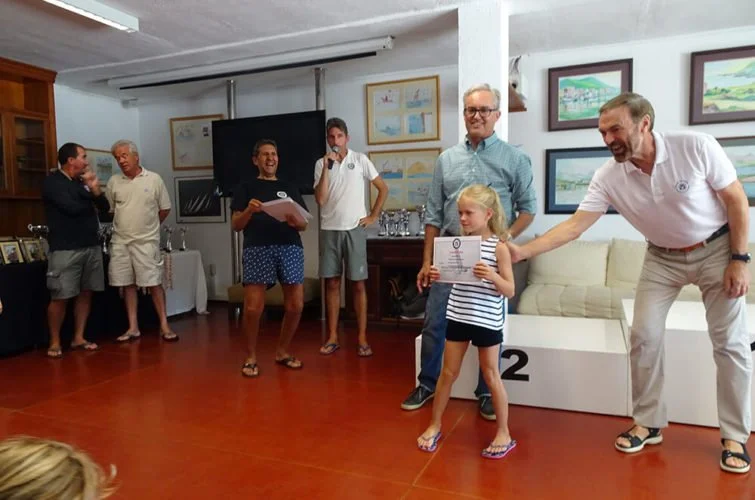 Niña recibiendo un certificado en una ceremonia en una sala con varias personas sonriendo y cuadros en las paredes.
