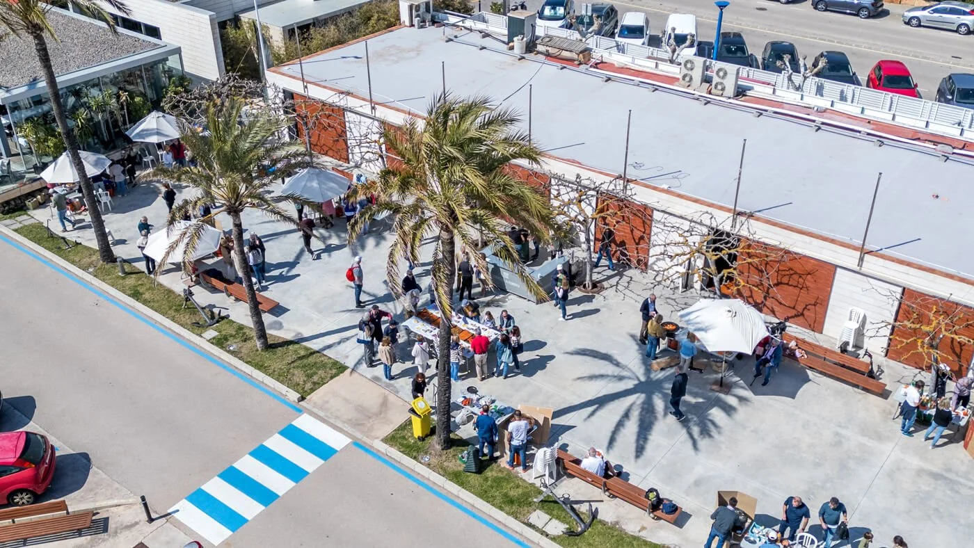 Vista aérea de un mercado callejero con varias mesas, sombrillas y personas comprando y vendiendo, árboles y un edificio con estacionamiento de fondo.