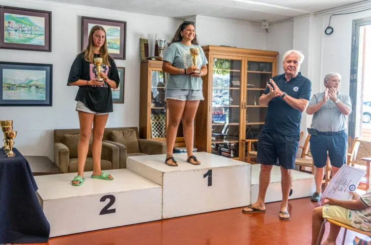 Mujeres en un podio recibiendo trofeos en una ceremonia, con una mujer en tercer lugar y otra en primer lugar, mientras una pareja de adultos los felicita.