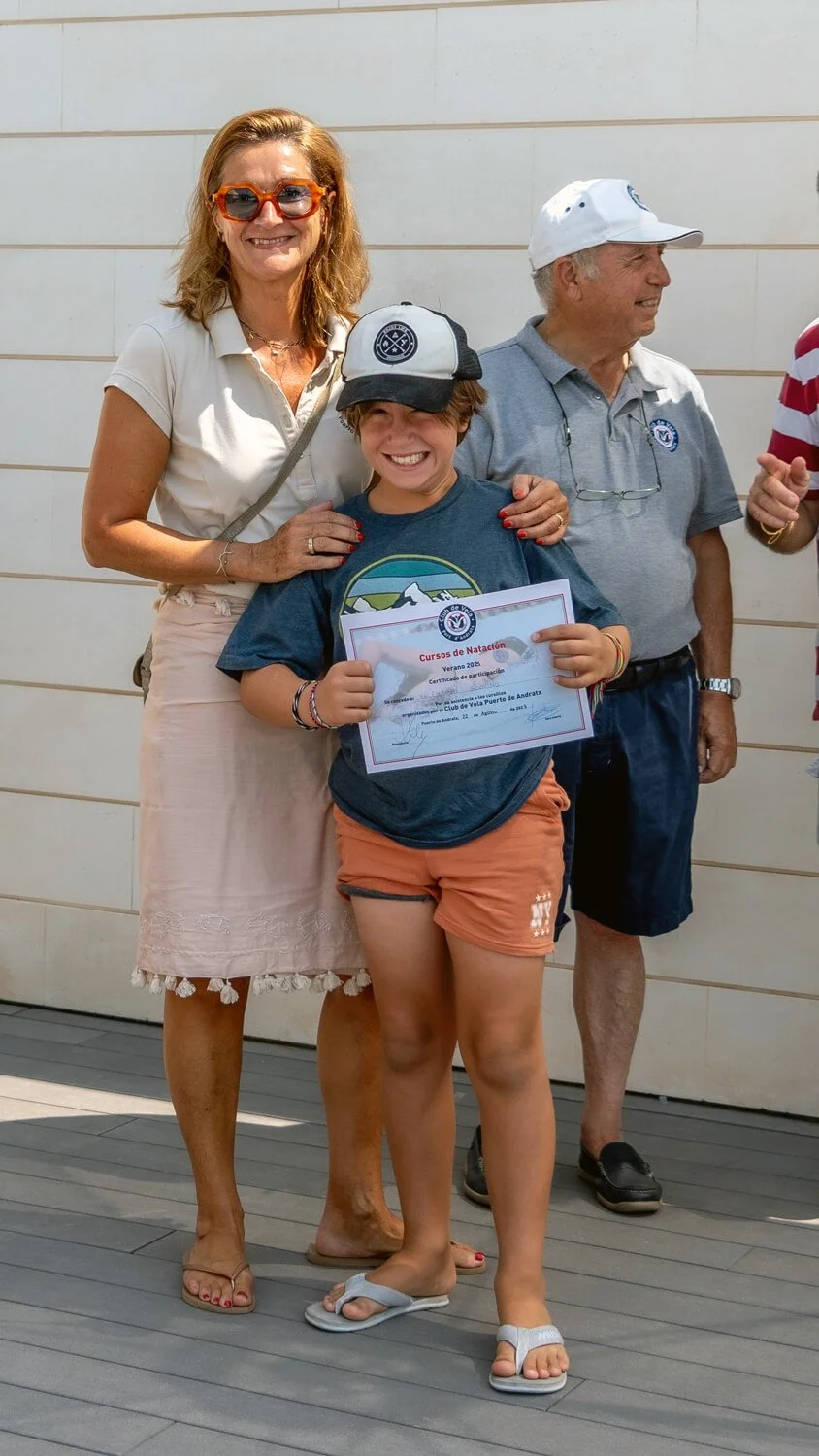 Niña sonriendo sosteniendo un certificado, acompañada por adultos en un evento de natación al aire libre.