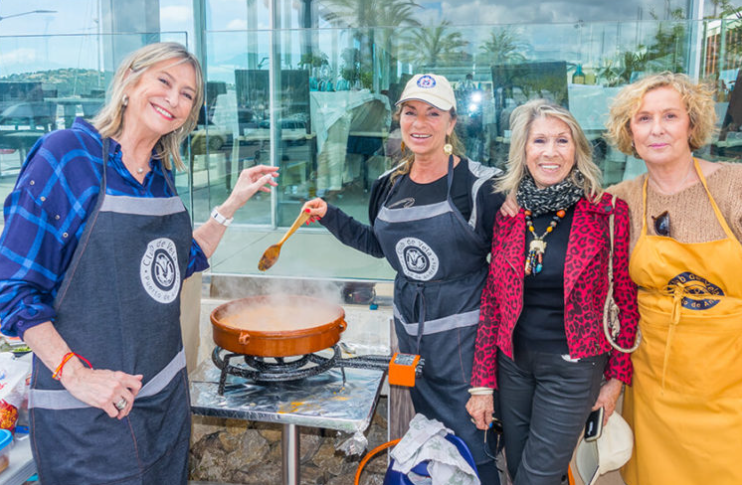 Cuatro mujeres cocinando juntas afuera, una de ellas mezclando en una olla grande, todas sonriendo y llevan delantales