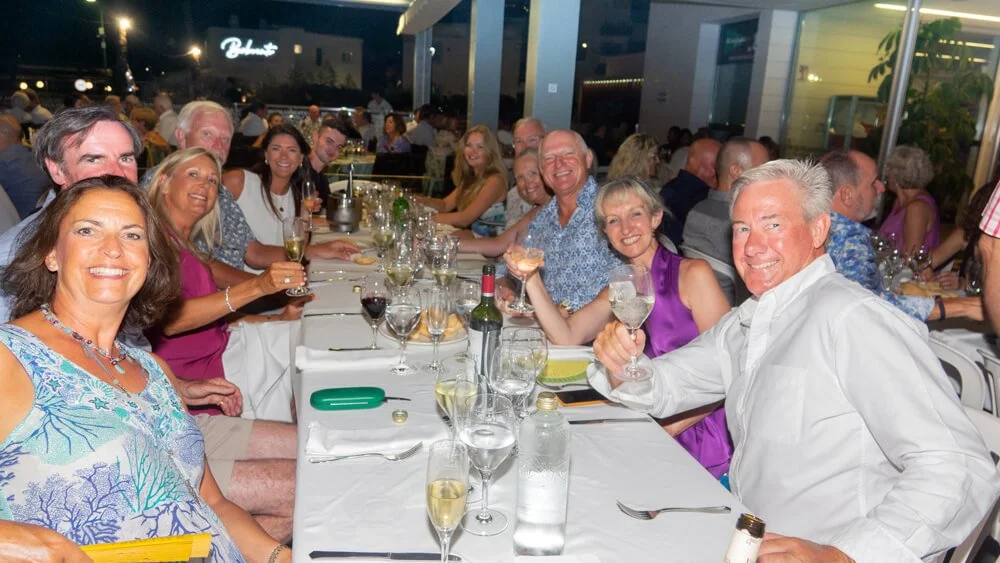 Un grupo de personas disfrutando de una comida en un restaurante, con varias mesas llenas de comida y bebidas, y un ambiente alegre en la noche.
