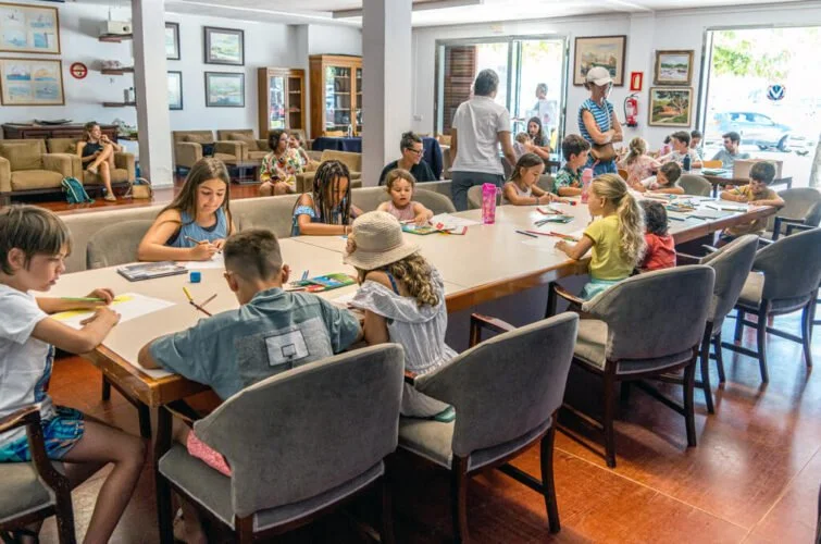 Niños pequeños participando en una actividad artística en un salón, acompañados por adultos, en un espacio con muchas ventanas y muebles de madera.