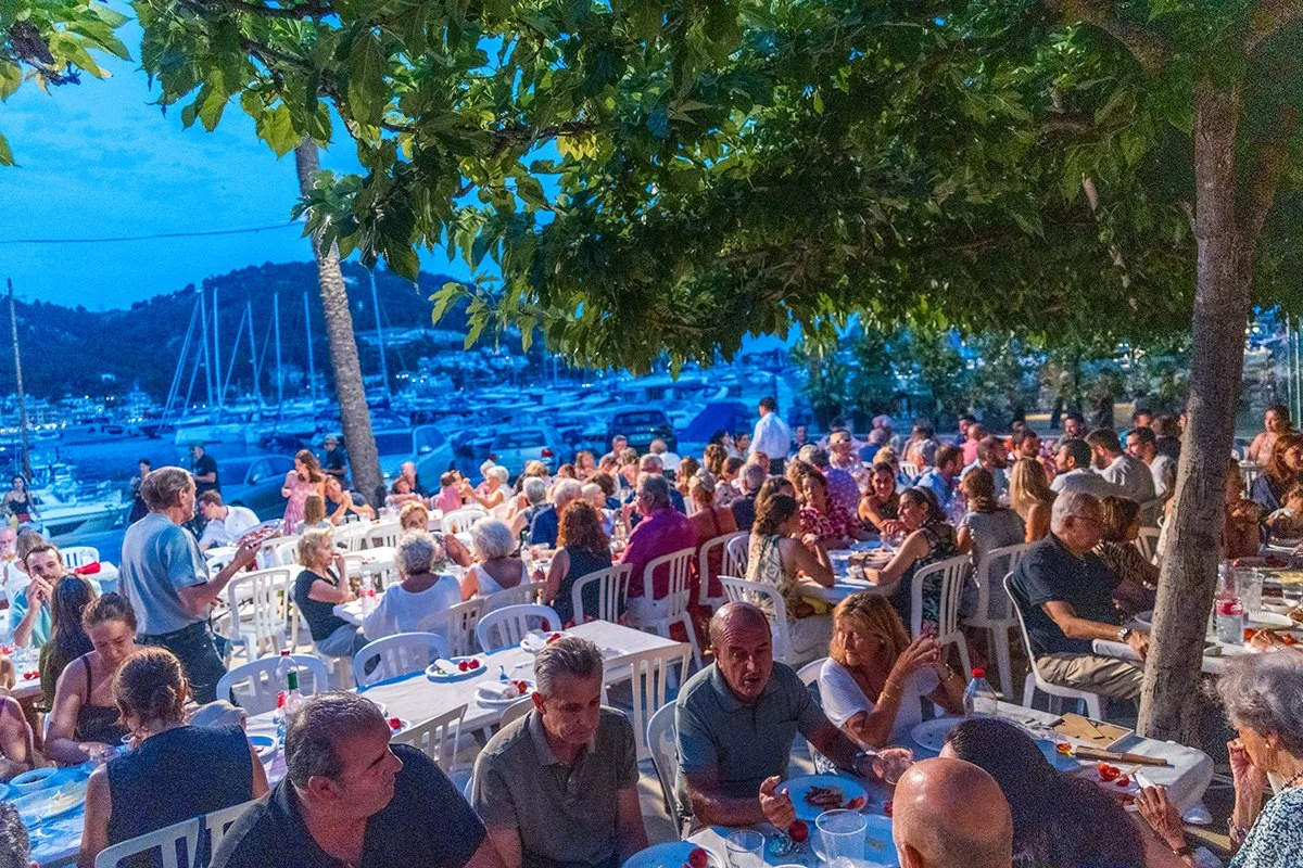 Una reunión social al aire libre con muchas mesas y personas disfrutando de una comida, en un entorno con árboles y vistas de un puerto con yates y montañas al fondo, en la noche.