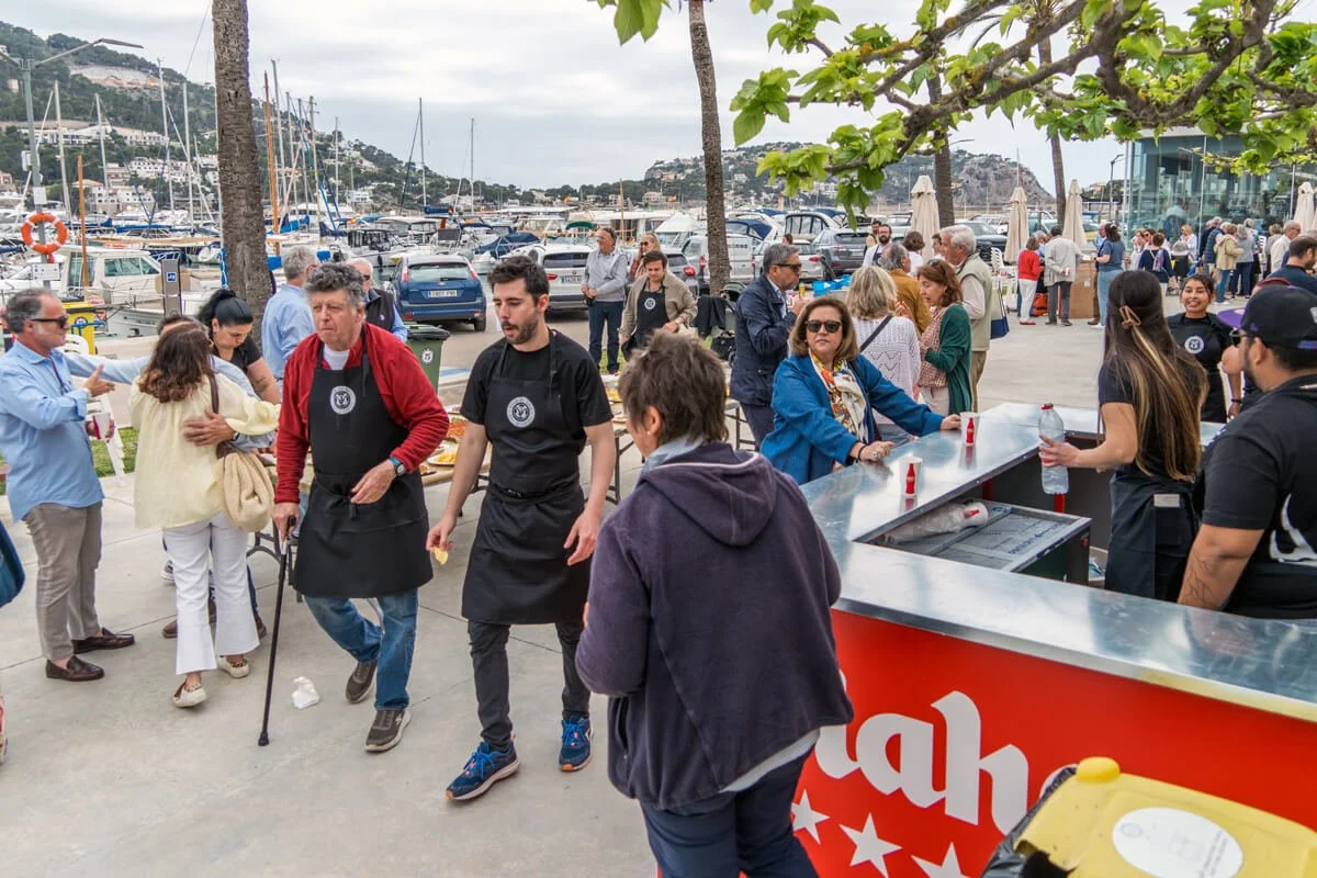 Personas en un puerto visitando puestos y disfrutando del día, con botes y yates en el fondo. Algunas personas están haciendo fila en un puesto de comida o bebidas.