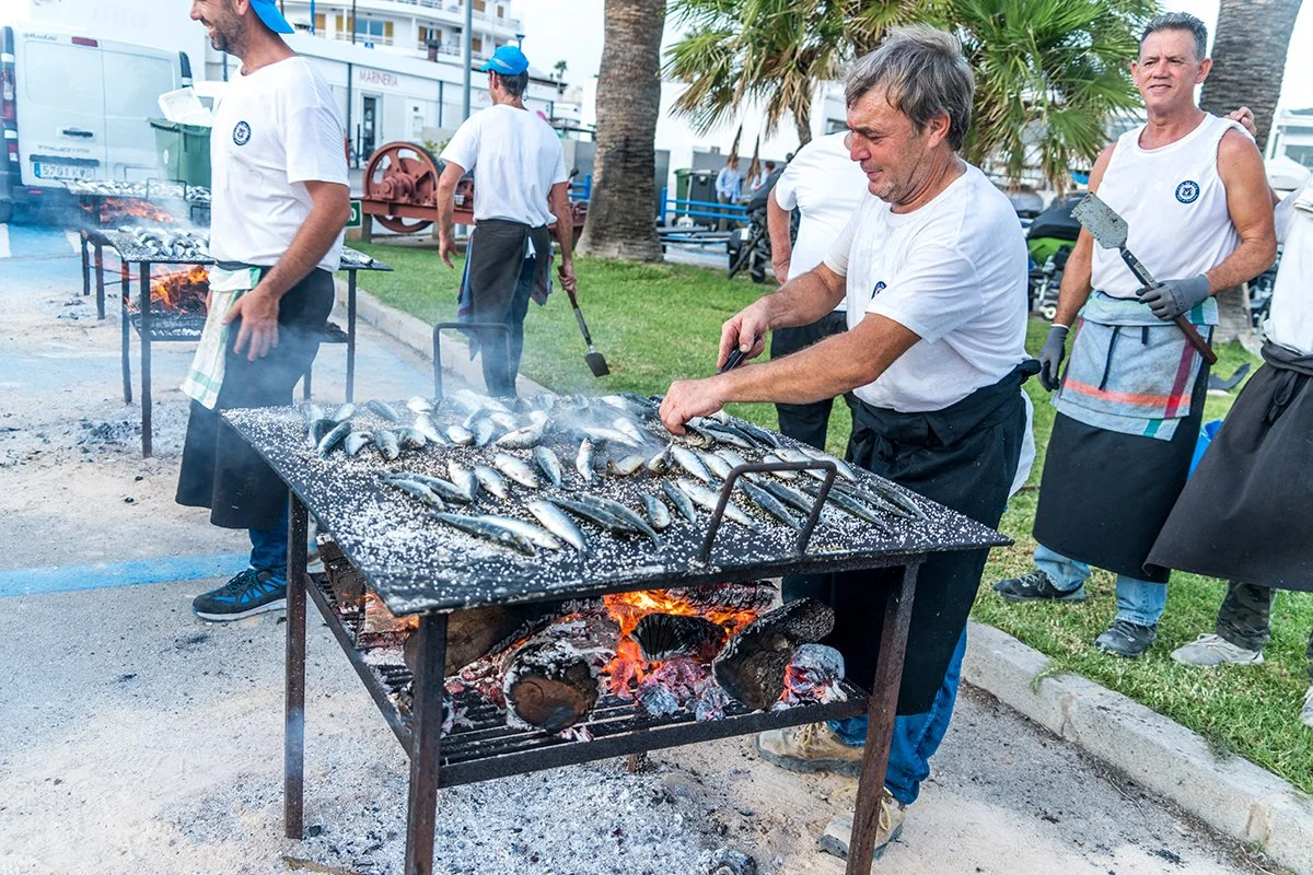 Hombres cocinando sardinas a la parrilla en un evento al aire libre en una zona con palmeras.