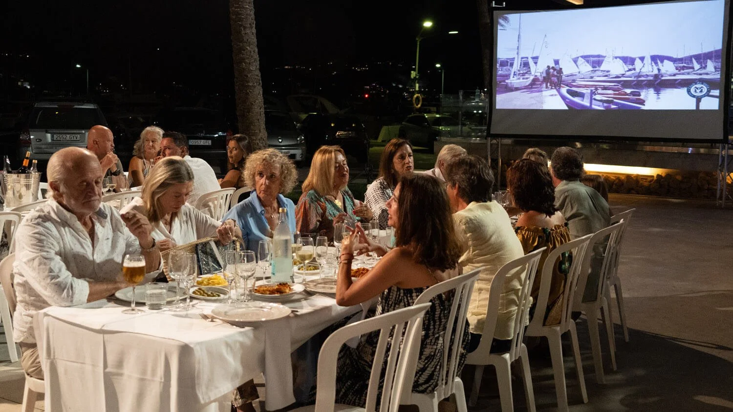 Cena de personas cenando en un restaurante al aire libre en la noche, con una pantalla grande que muestra una imagen de barcos en un puerto.