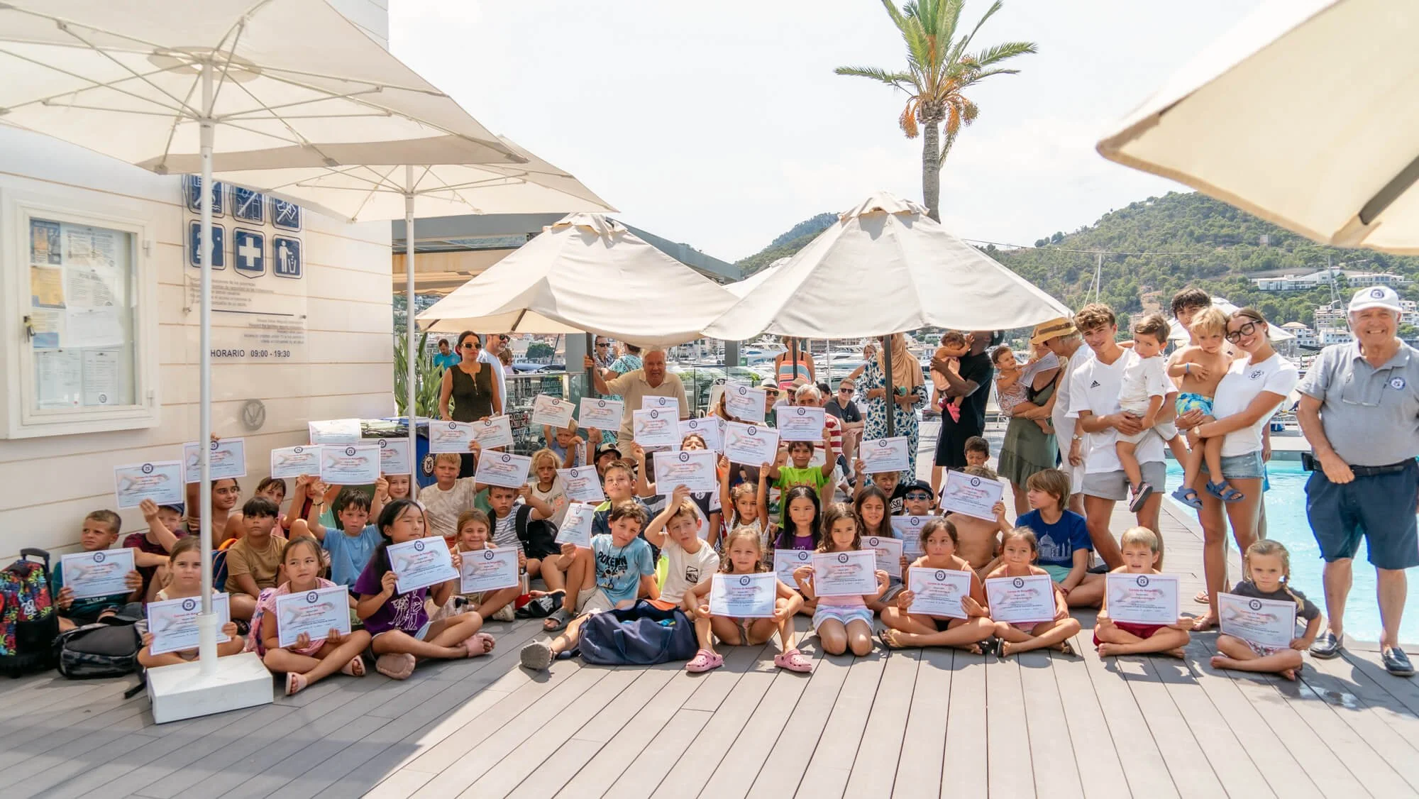 Grupo de niños y adultos posando con diplomas en un evento al aire libre junto a una zona de piscinas, con sombrillas y árboles en el fondo.