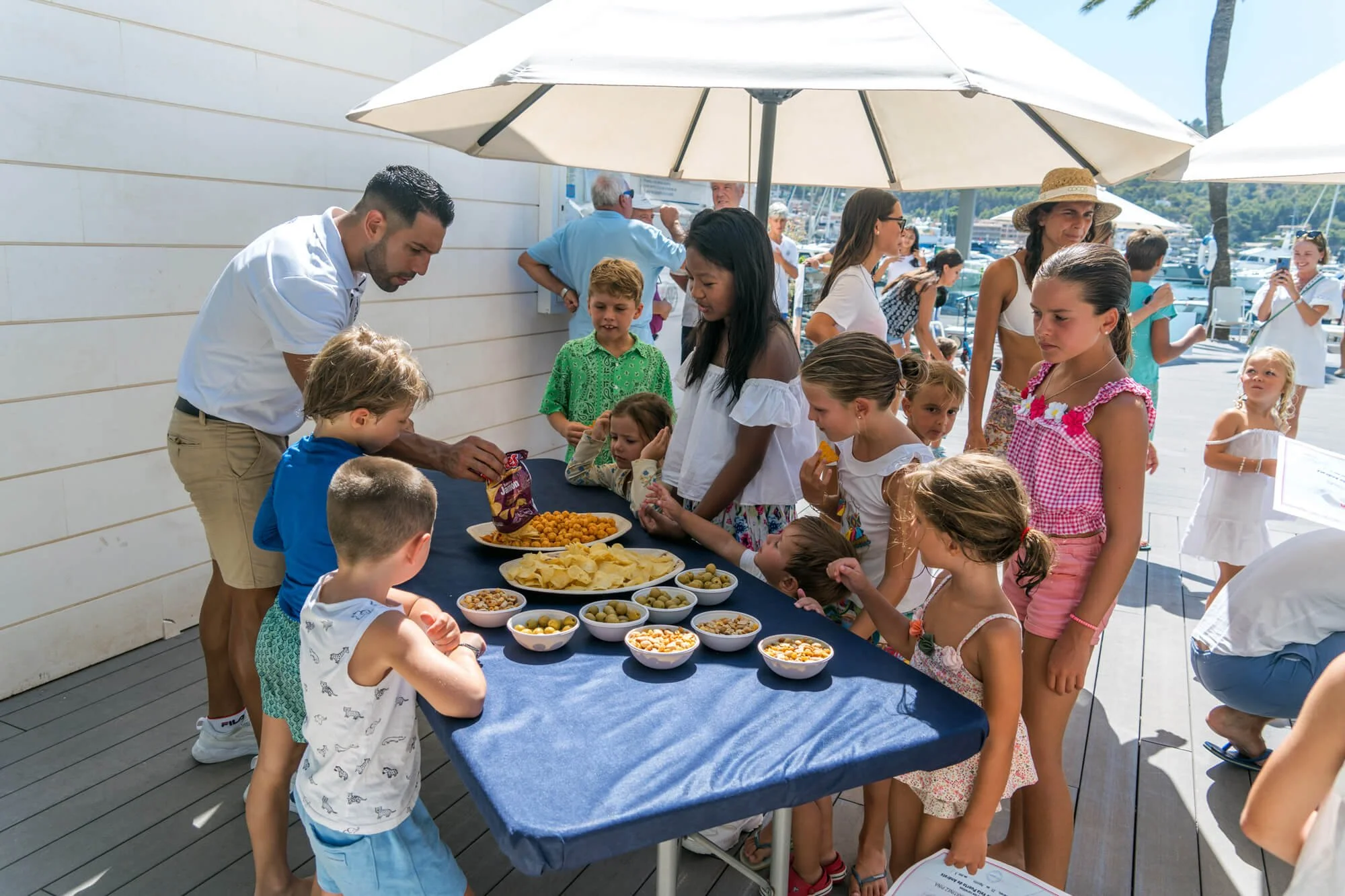Niños y adultos en una fiesta en la terraza, sirviéndose comida y disfrutando del día en un evento al aire libre cerca del mar.