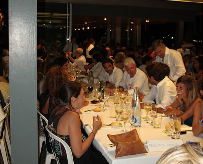 Grupo de personas en una cena elegante, sentadas alrededor de una mesa larga con comida y bebidas, en un ambiente formal y festivo.