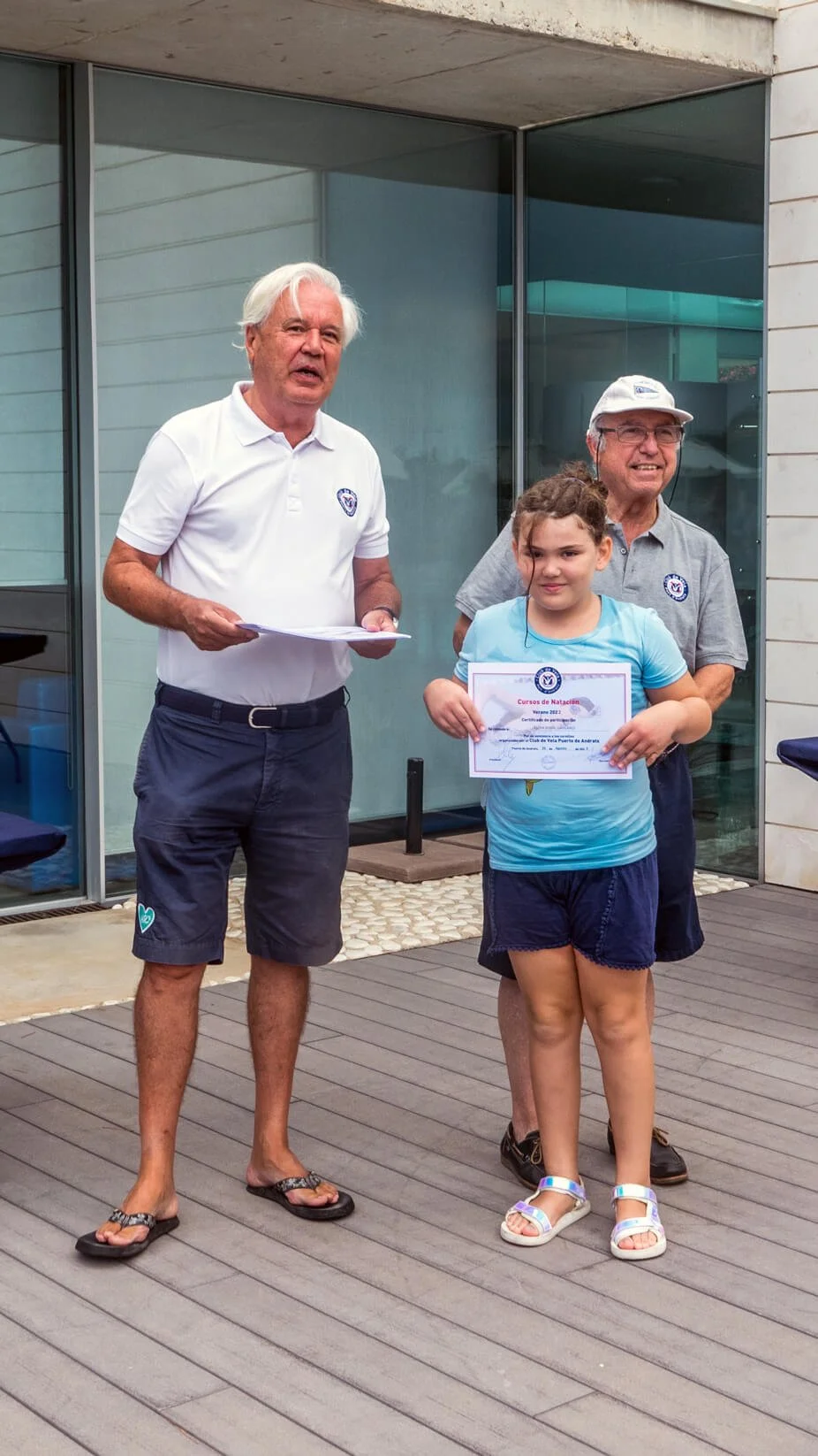 Una niña recibe un certificado de natación de manos de un hombre mayor en un evento al aire libre, acompañado de dos adultos masculinos, en un entorno moderno con ventanas de vidrio y piso de madera.