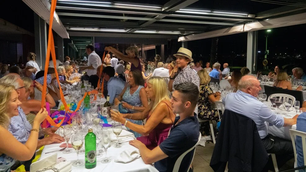 Personas en una cena en un lugar cerrado, con mesas largas, muchas copas y botellas, algunos invitados vestidas elegantemente, ambiente festivo y social.